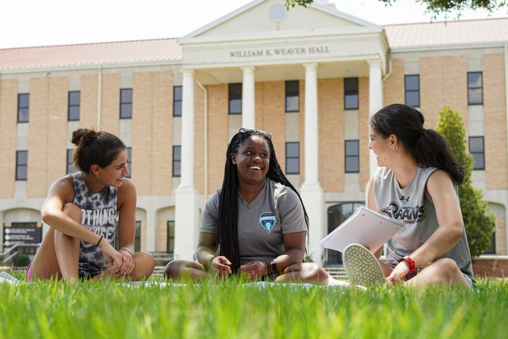 three college girls sitting on the grass in front of a building on campus.